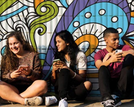 Cinq adolescents clavardent sur leur téléphone, devant un mur avec un dessin