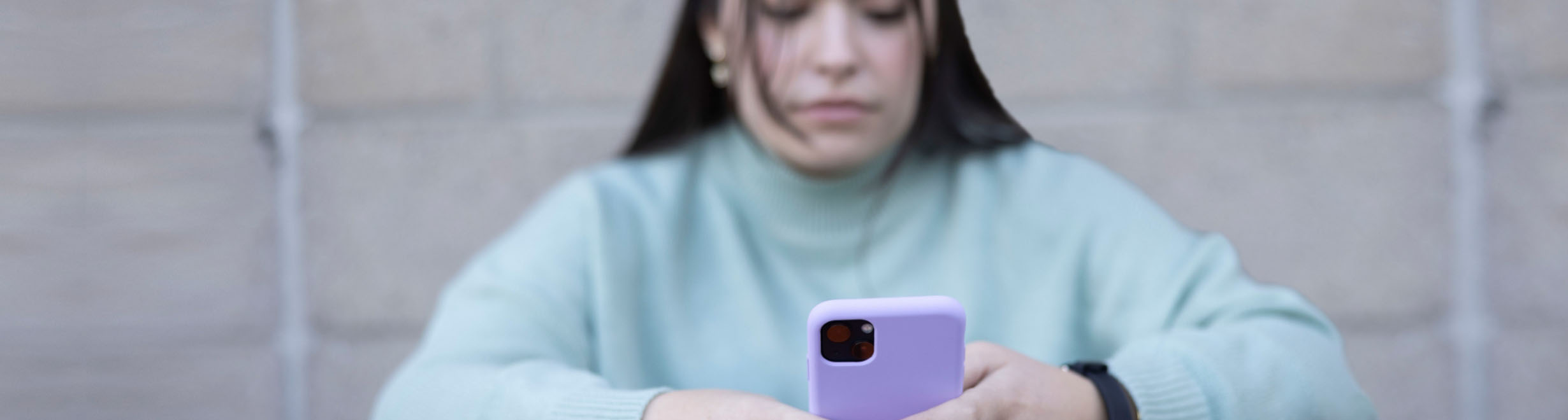 Jeune personne portant un gilet vert assise devant un mur de briques et utilisant le service de clavardage en direct sur un téléphone intelligent violet